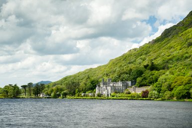 Ein majestätisches Schloss liegt am Ufer eines ruhigen Sees, umgeben von üppigem, grünem Wald und sanften Hügeln.