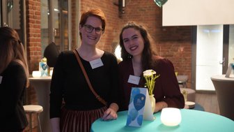 Zwei Frauen lächeln an einem Tisch mit einer blauen Tischdecke, umgeben von einer festlichen Atmosphäre mit Lichtern.