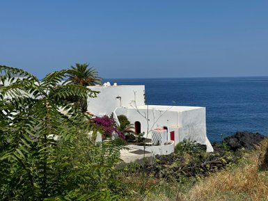 Ein Blick auf eine weiße Villa mit roten Akzenten, umgeben von üppigem Grün und dem blauen Meer im Hintergrund.