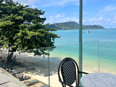 Ein Blick auf den Strand von einem Balkon mit einem Tisch und einem Stuhl, umgeben von Palmen und klarem Wasser.