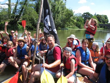 Eine fröhliche Gruppe von Kindern und Erwachsenen auf einem Kanu, umgeben von Natur und Sonnenschein, genießt den Tag.