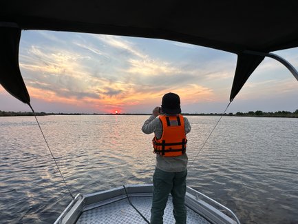Ein Mensch in einer orangefarbenen Schwimmweste fotografiert den Sonnenuntergang über einem ruhigen Gewässer.