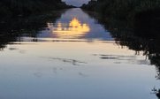 Ein ruhiger Wasserweg spiegelt die warmen Farben eines Sonnenuntergangs wider, umgeben von üppigem Grün und sanften Wolken.