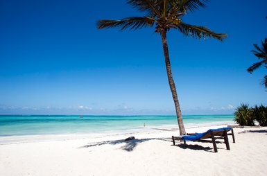 Ein einsamer Liegestuhl steht am weißen Sandstrand, umgeben von klarem, türkisfarbenem Wasser und einem strahlend blauen Himmel.