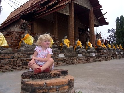 Ein kleines Mädchen mit blonden Locken sitzt auf einem Ziegelstein und schaut lächelnd zu den Buddha-Statuen im Hintergrund.