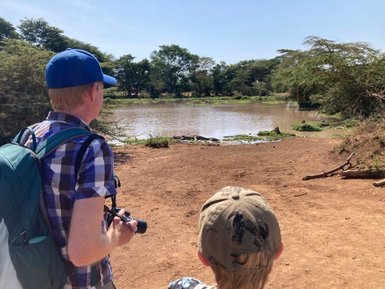 Zwei Personen stehen am Ufer eines ruhigen Gewässers, umgeben von üppiger Vegetation und einem klaren blauen Himmel.
