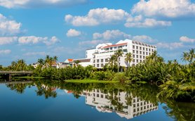 Ein modernes Hotelgebäude spiegelt sich im ruhigen Wasser eines Teiches, umgeben von üppigem Grün und Palmen.