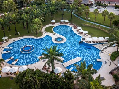 Ein weitläufiger Pool mit blauen Fliesen, umgeben von Palmen und Liegen, lädt zum Entspannen in der Sonne ein.