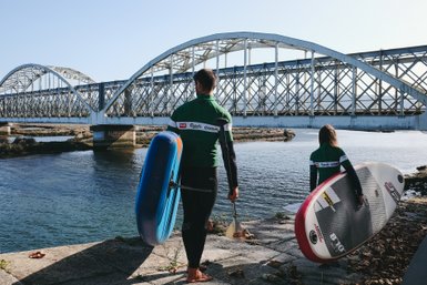Ein Mann und ein Kind stehen am Wasser, bereit zum Paddeln, während eine Brücke im Hintergrund sichtbar ist.
