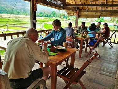 Eine Gruppe von Menschen sitzt an Holztischen und genießt eine Mahlzeit in einem offenen Restaurant mit Blick auf Reisfelder.