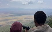 Zwei Personen stehen an einem Aussichtspunkt und betrachten mit einem Fernglas die weite Landschaft unter einem bewölkten Himmel.