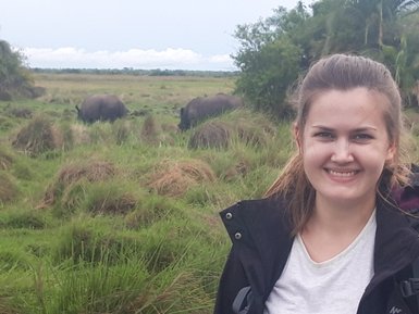 Eine lächelnde Frau steht in einer grünen Landschaft, während im Hintergrund zwei Nashörner grasen.