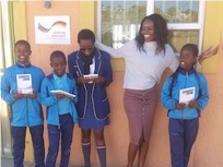 Fünf Kinder stehen vor einem Gebäude, lächeln und halten Bücher in den Händen, während eine Frau neben ihnen posiert.