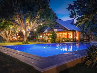 Ein glänzender, blauer Pool vor einem gemütlichen Restaurant, umgeben von großen Bäumen mit bunten Lichtern.