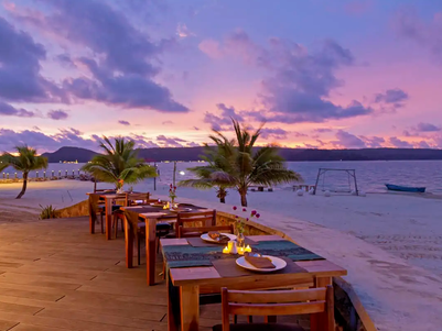 Ein romantisches Restaurant am Strand mit Holztischen, umgeben von Palmen und einem atemberaubenden Sonnenuntergang im Hintergrund.
