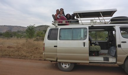 Eine Gruppe von Kindern und Erwachsenen sitzt fröhlich auf dem Dach eines Vans, umgeben von einer ländlichen Landschaft.