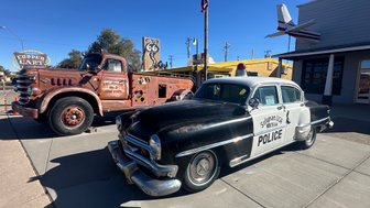 Ein klassisches Polizeiauto steht neben einem alten, rostigen Lastwagen in einer sonnigen Stadtlandschaft.