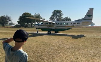 Ein Junge mit einer Kappe beobachtet ein kleines Flugzeug auf einem offenen Feld in Botswana unter klarem Himmel.