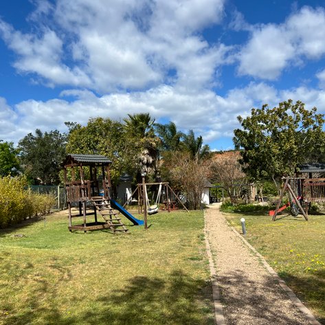 Ein Spielplatz im Freien mit Holzspielgeräten, umgeben von üppigem Grün und einem klaren blauen Himmel.