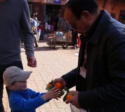 Ein fröhlicher Junge in blauer Jacke reicht einem Verkäufer eine Orange, während ein kleiner Junge im Kinderwagen zuschaut.