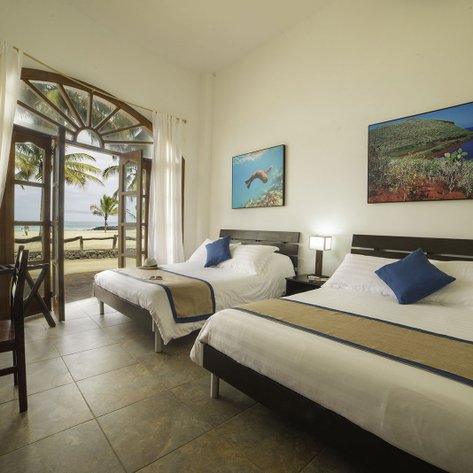 Ein helles Zimmer mit zwei Betten, Blick auf den Strand und Palmen durch große Fenster, die eine entspannende Atmosphäre schaffen.