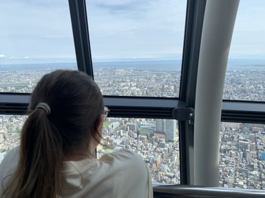 Eine Person mit einem Pferdeschwanz blickt aus dem Fenster des Tokio Skytree auf die weitläufige Stadtlandschaft.