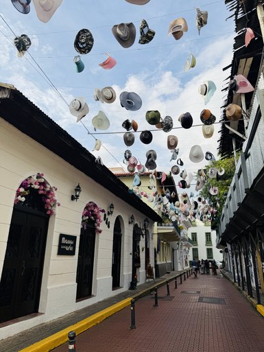 Bunte Hüte hängen über einer malerischen Straße in Panama, umgeben von historischen Gebäuden und blühenden Pflanzen.
