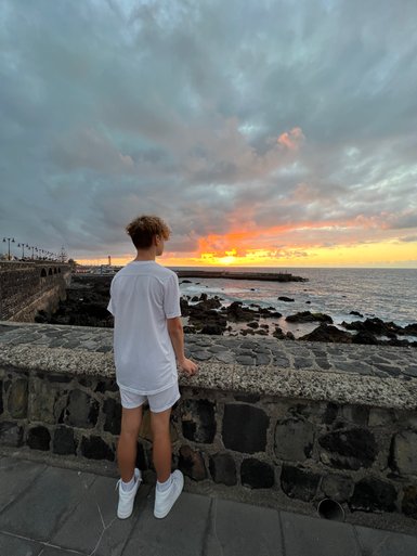 Ein junger Mann in weißer Kleidung steht an einer Steinmauer und betrachtet den Sonnenuntergang über dem Meer.