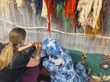 Zwei Frauen arbeiten an einem Teppich, umgeben von bunten Wollsträngen und einem netten, handgefertigten Teppich.