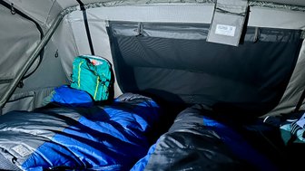 Im Inneren eines Zeltes liegen zwei Schlafsäcke in Blau und Grau, während ein grüner Rucksack in der Ecke steht.