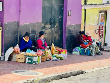 Drei Frauen in traditioneller Kleidung verkaufen frisches Obst und Gemüse auf der Straße vor bunten Wänden.