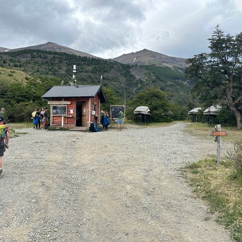 Ein Wanderer mit Rucksack geht auf einem Schotterweg zu einer kleinen roten Hütte in der Natur Patagoniens.