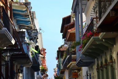 Bunte Balkone mit Blumen schmücken die engen Gassen einer historischen Stadt, unter einem strahlend blauen Himmel.