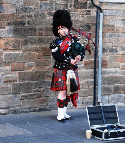 Ein schottischer Dudelsackspieler in traditioneller Kleidung steht vor einer Steinmauer und spielt leidenschaftlich seine Musik.