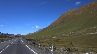 Eine kurvenreiche Straße schlängelt sich durch eine grüne Landschaft, umgeben von majestätischen Bergen unter einem klaren blauen Himmel.