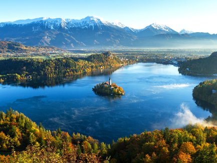 Ein malerischer Blick auf den Bleder See, umgeben von buntem Herbstlaub und majestätischen Bergen im Hintergrund.