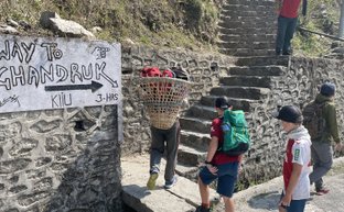 Eine Gruppe von Kindern steht an einer steinernen Treppe, während ein Erwachsener mit einem Korb auf dem Rücken vorbeigeht.