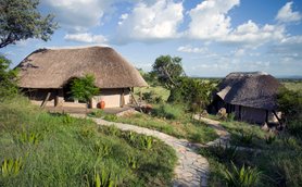 Zwei strohgedeckte Hütten stehen in einer grünen Landschaft, umgeben von Bäumen und sanften Hügeln unter einem blauen Himmel.
