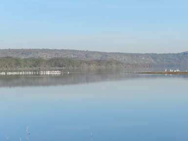 Ein ruhiger See mit einer spiegelglatten Oberfläche, umgeben von sanften Hügeln und einer Gruppe von Flamingos im Hintergrund.