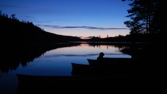 Ein ruhiger See bei Dämmerung, mit einem Silhouettenbild eines Mannes in einem Boot, umgeben von Bäumen.