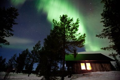 Ein Holzhaus steht in einer verschneiten Landschaft, während die Nordlichter in leuchtendem Grün darüber tanzen.