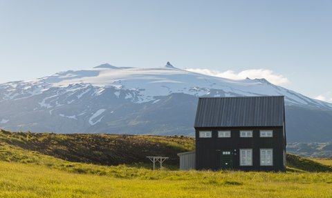Ein schwarzes Sommerhaus steht in einer grünen Wiese, mit schneebedeckten Bergen im Hintergrund, die unter blauem Himmel leuchten.