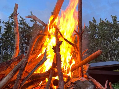Ein loderndes Lagerfeuer aus Ästen und Holzscheiten, umgeben von einer ruhigen, bewaldeten Landschaft unter einem blauen Himmel.