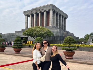 Drei Frauen posieren fröhlich vor dem Ho-Chi-Minh-Mausoleum, umgeben von gepflegten Pflanzen und einem klaren Himmel.