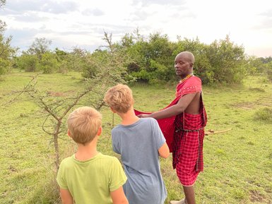 Ein Massai erklärt Kindern die Natur während eines Spaziergangs in der afrikanischen Savanne, umgeben von grünen Büschen.