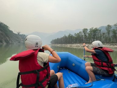 Zwei Kinder in roten Schwimmwesten paddeln in einem blauen Schlauchboot auf einem ruhigen Fluss in Nepal.