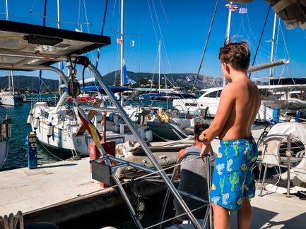 Ein Junge mit nacktem Oberkörper steht an einem Boot und beobachtet die anderen Yachten im Hafen unter strahlend blauem Himmel.