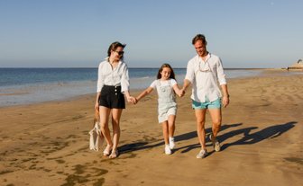 Eine Familie spaziert fröhlich am Strand entlang, während die Sonne am Himmel scheint und das Wasser sanft plätschert.