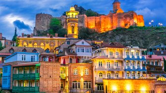 Die malerische Altstadt von Tiflis zeigt bunte Häuser und eine beeindruckende Festung im Hintergrund, beleuchtet bei Dämmerung.