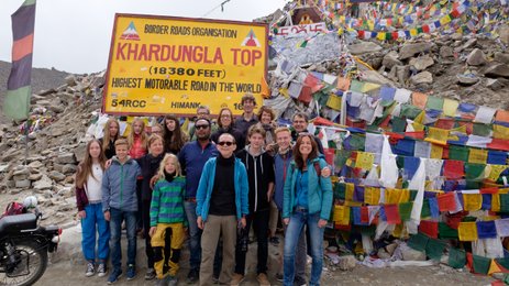 Eine Gruppe von Reisenden posiert fröhlich vor dem Khardungla-Pass-Schild, umgeben von bunten Gebetsfahnen.